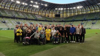 Grupa ludzi na murawie stadionu piłkarskiego. W centrum zdjęcia stoi i siedzi kilkadziesiąt osób – są zarówno mężczyźni, kobiety, jak i dzieci, w tym kilka osób na wózkach inwalidzkich. Część osób ubrana jest w żółte koszulki, a jedna kobieta z przodu trzyma żółtą koszulkę piłkarską z napisem „BŁĄD” i numerem 11. W tle widać puste trybuny stadionu oraz charakterystyczny dach z łukami i oświetleniem.