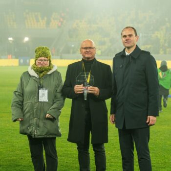 3 osoby stoją na murawie stadionu piłkarskiego. Osoba po lewej ma zieloną, zimową kurtkę, zielono-żółtą czapkę i szalik, okulary oraz duży identyfikator na szyi. Osoba w środku jest ubrana na czarno i ma okulary, a w rękach trzyma przezroczystą statuetkę o kształcie sześciokąta. Osoba po prawej ubrana jest w ciemny płaszcz. W tle widać trybuny stadionu z żółtymi siedzeniami w lekkiej mgle i kilka osób na murawie.